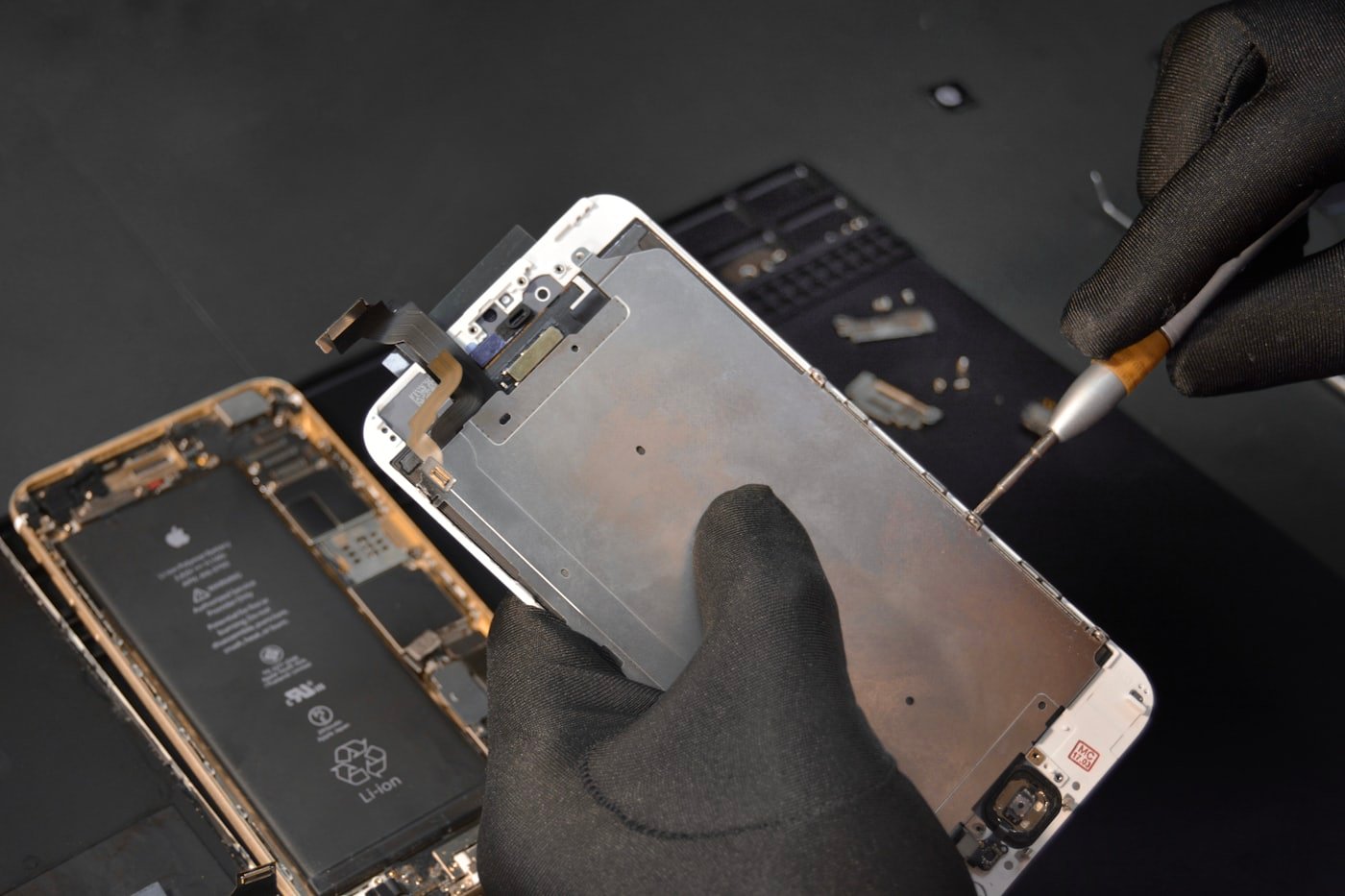 Técnico de luvas removendo o display de um iPhone com chave de precisão, com a bateria original visível ao lado
